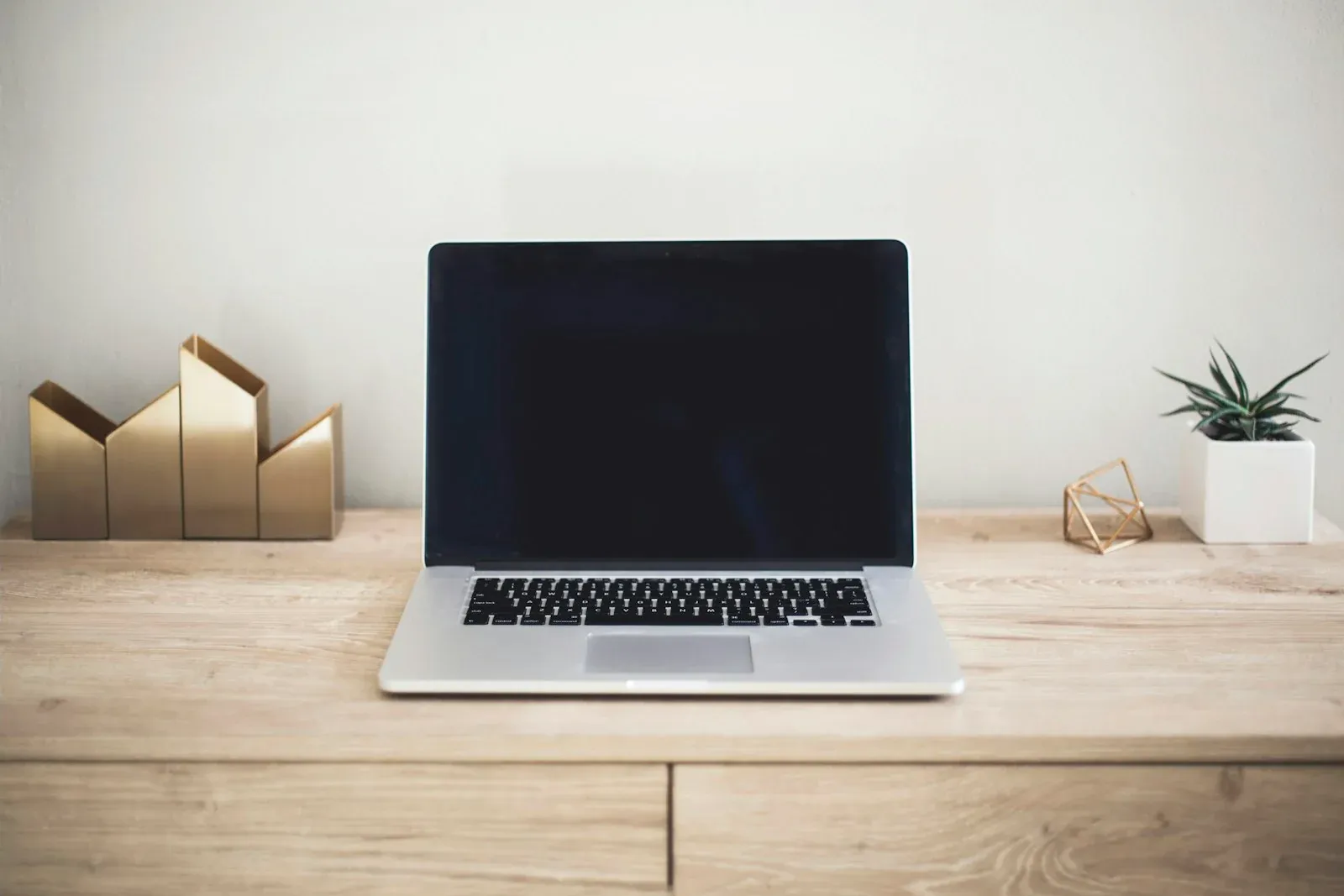 An open laptop on a wooden desk beside a notebook and small plant